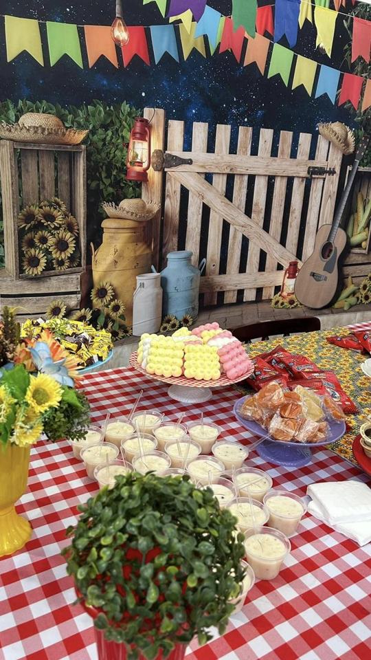Mesa da festa junina com toalha xadrez vermelha, doces, flores e um cenário de porteira e sanfona